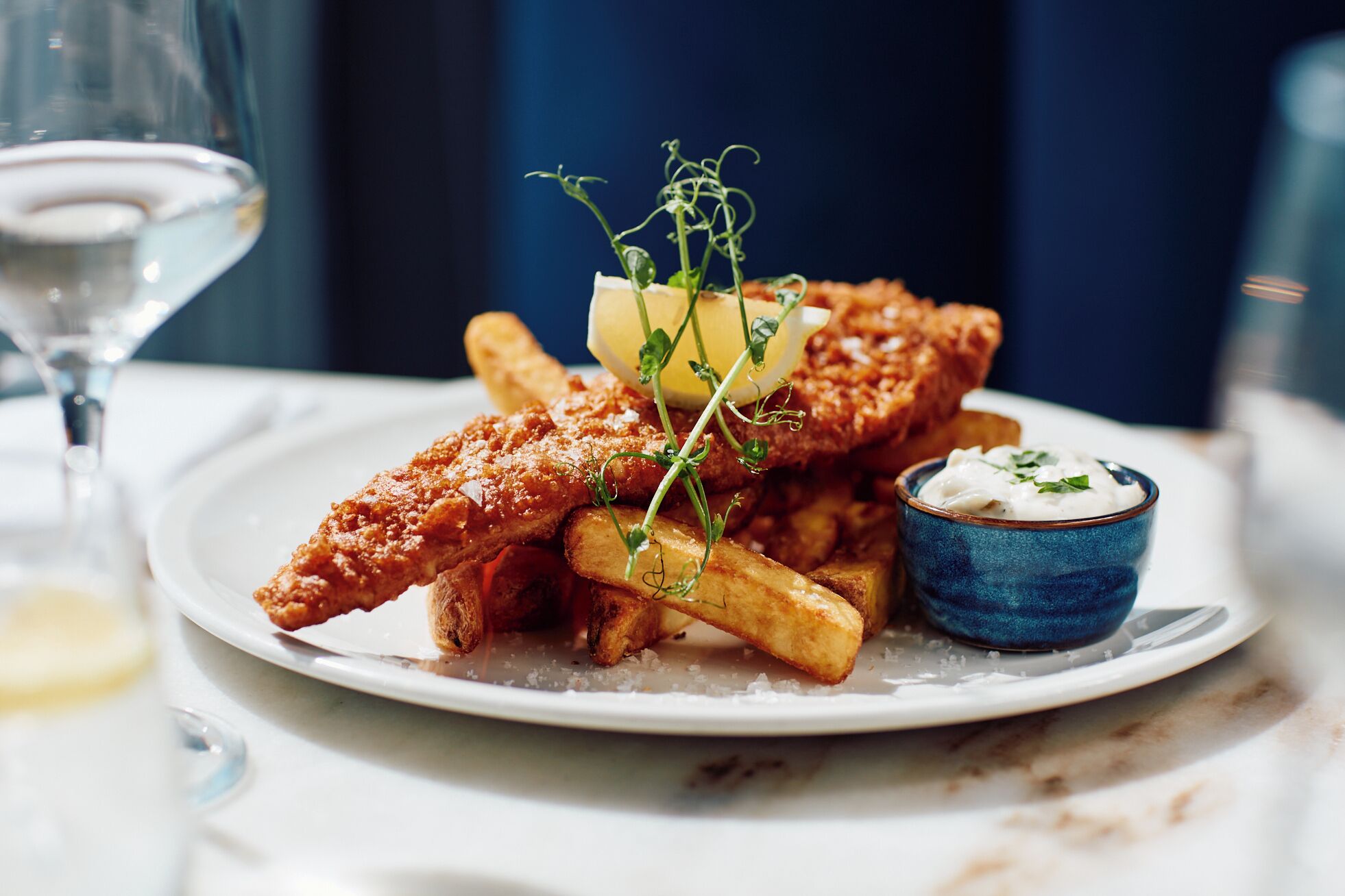 A plate of fried fish, fries, and a small bowl of sauce, with a glass of white wine in the background.