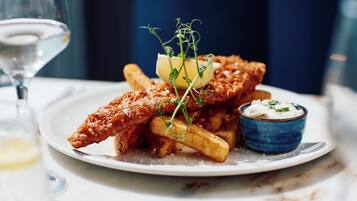 A plate of fried fish, fries, and a small bowl of sauce, with a glass of white wine in the background.