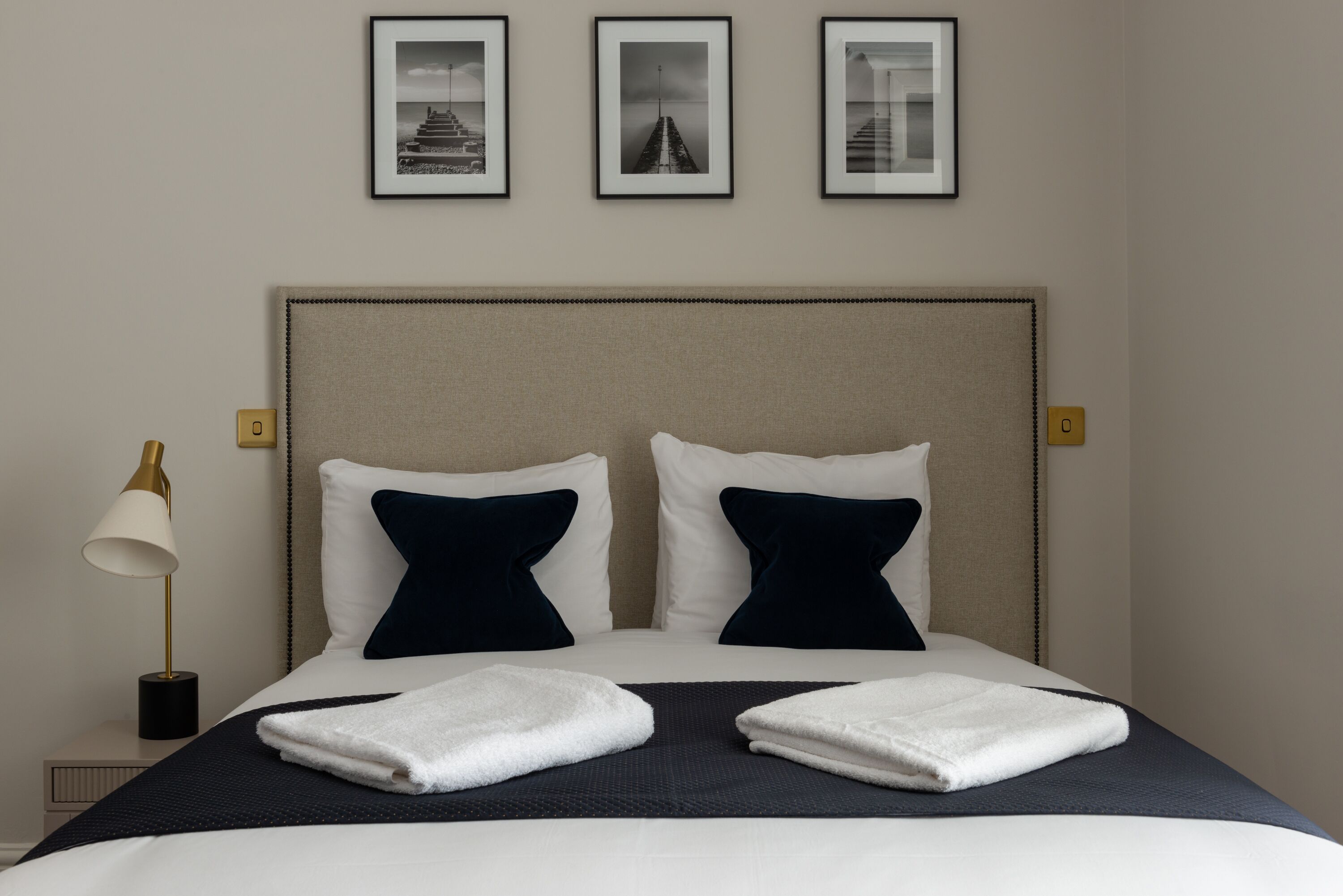Una cama bien hecha con almohadas blancas y azul marino, una colcha oscura y dos toallas blancas dobladas. Encima de la cama, hay tres cuadros enmarcados en la pared.