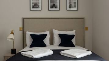 Una cama bien hecha con almohadas blancas y azul marino, una colcha oscura y dos toallas blancas dobladas. Encima de la cama, hay tres cuadros enmarcados en la pared.