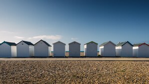 A row of beach huts with a gravel beach in the foreground and a clear blue sky. - The Burlington Hotel (Worthing)