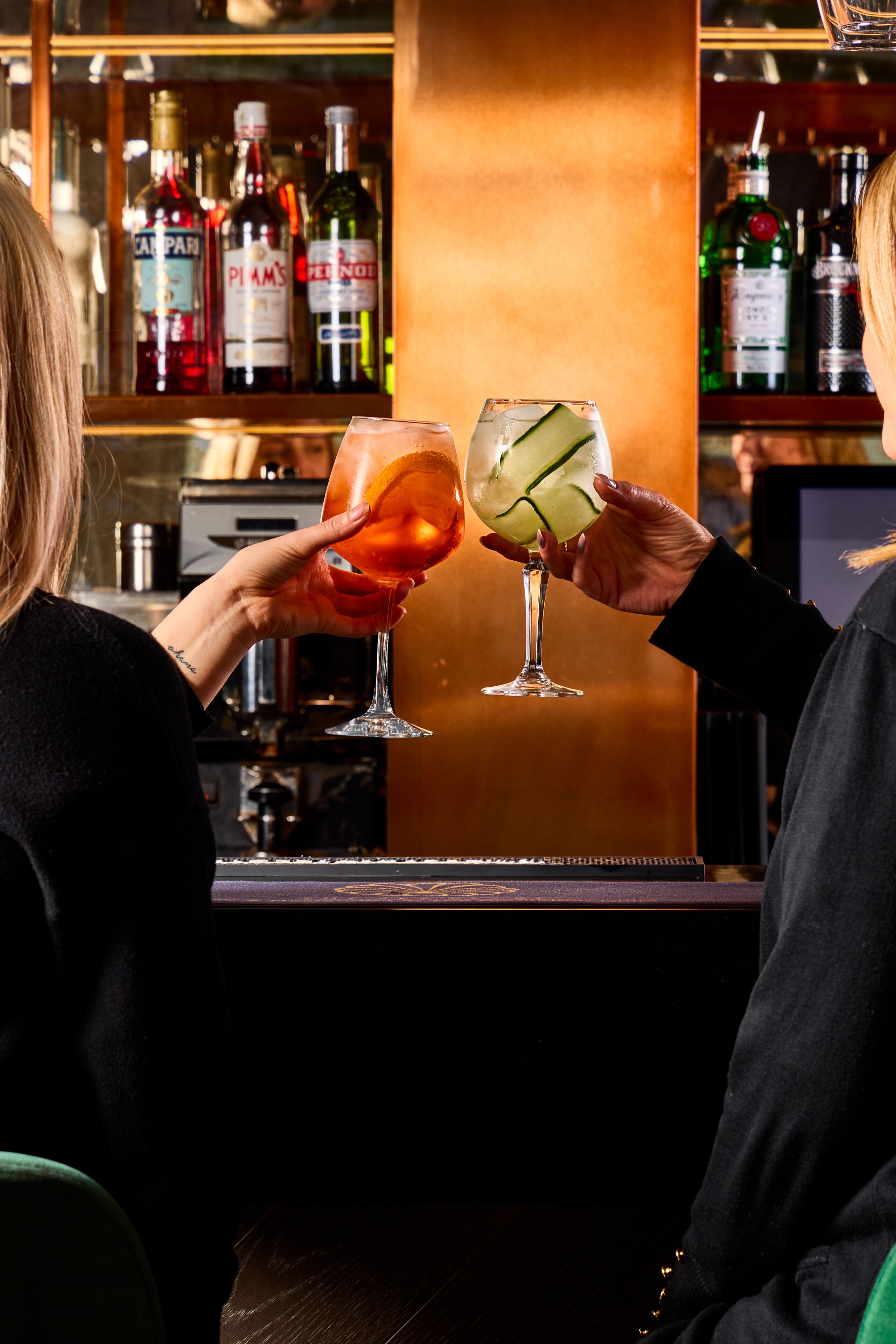 A bar with two people holding drinks. There are bottles of liquor on shelves behind them.