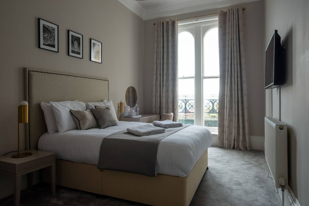 A hotel bedroom with a large bed, a bedside table, a mirror, and a view of the outside through a window.