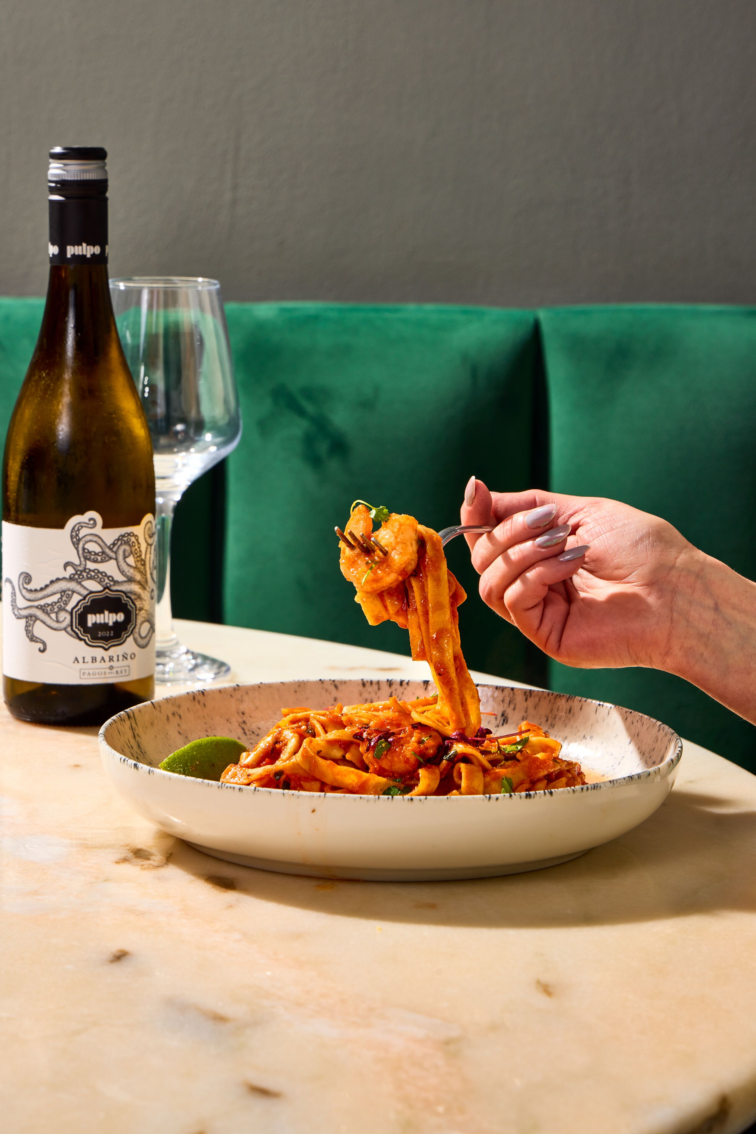 A plate of spaghetti with a fork lifting a portion, a wine bottle, and a glass on a table.