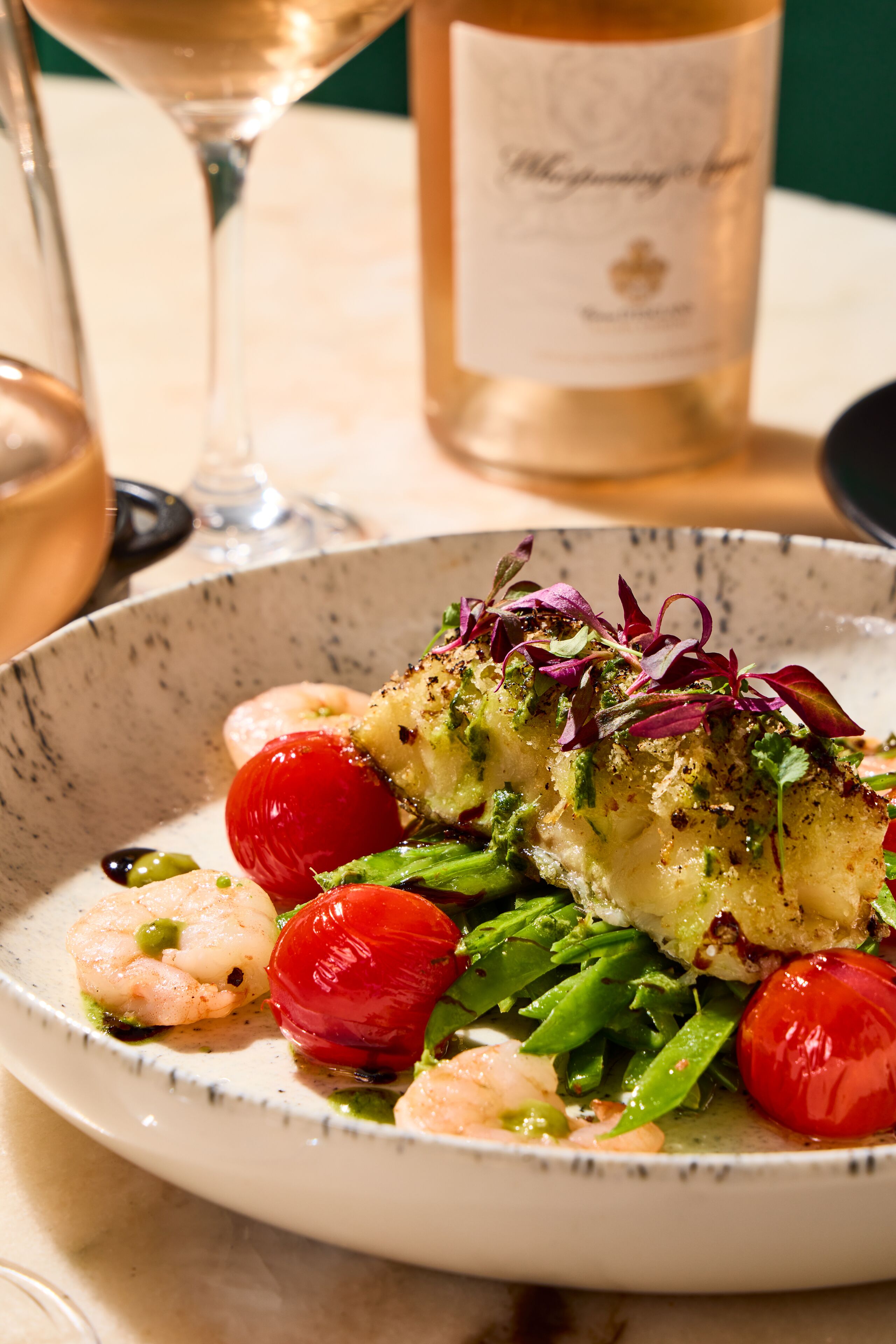 Une assiette de nourriture comprenant des crevettes, des tomates cerises et des légumes verts, avec une bouteille de vin en arrière-plan.