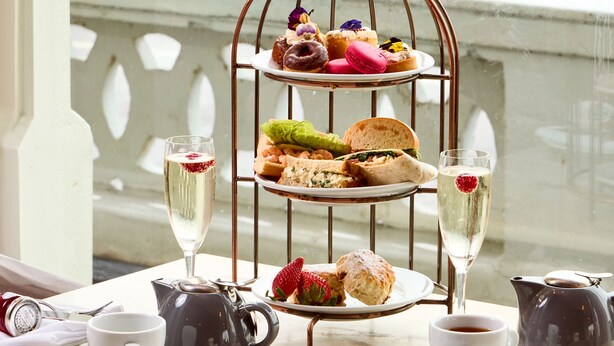 A three-tiered dessert stand with various pastries and sandwiches, a teapot, cups, and a view of a beach and bench in the background. - The Burlington Hotel (Worthing)