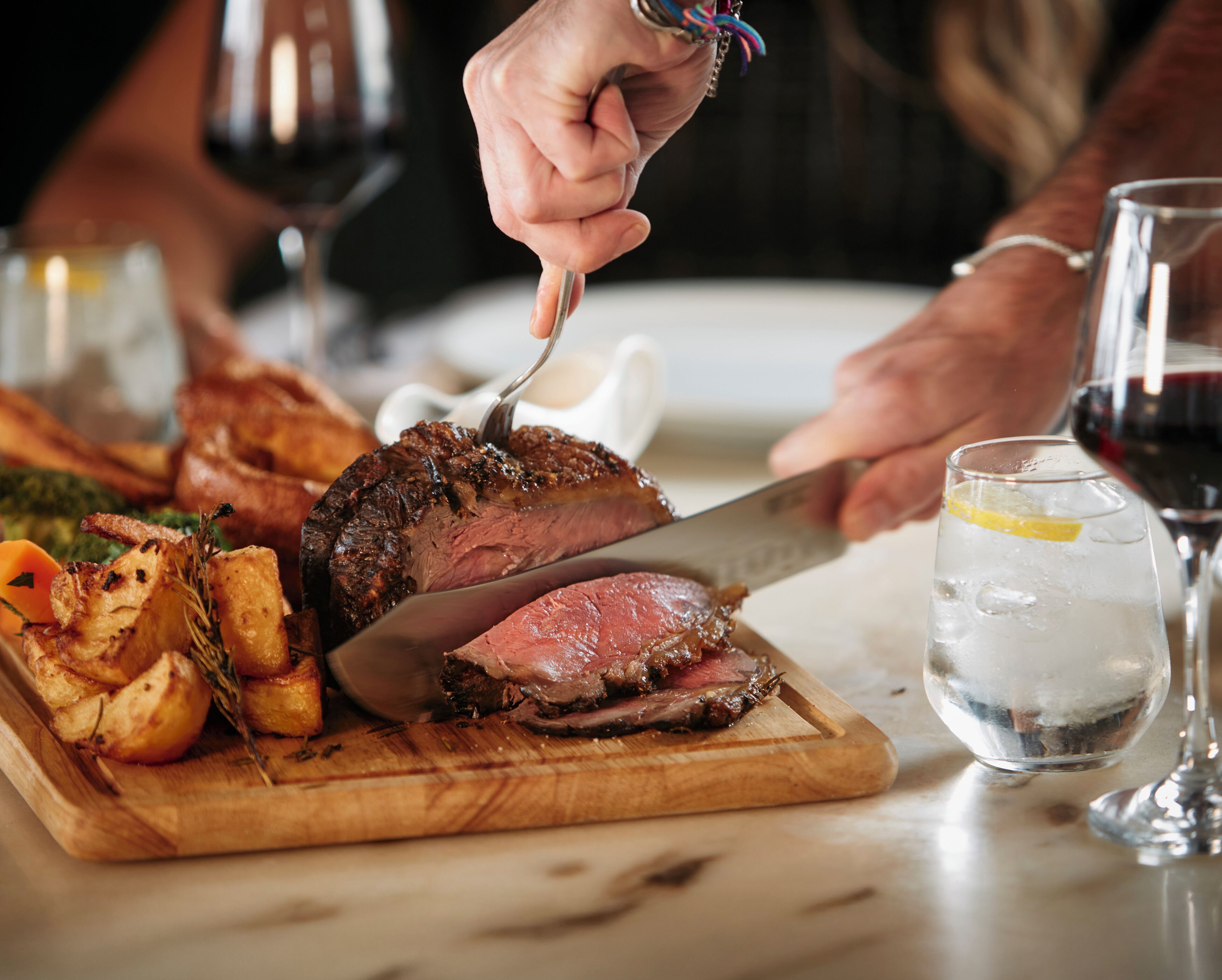 A person cutting a steak on a wooden cutting board with a knife, surrounded by food and a glass of red wine.