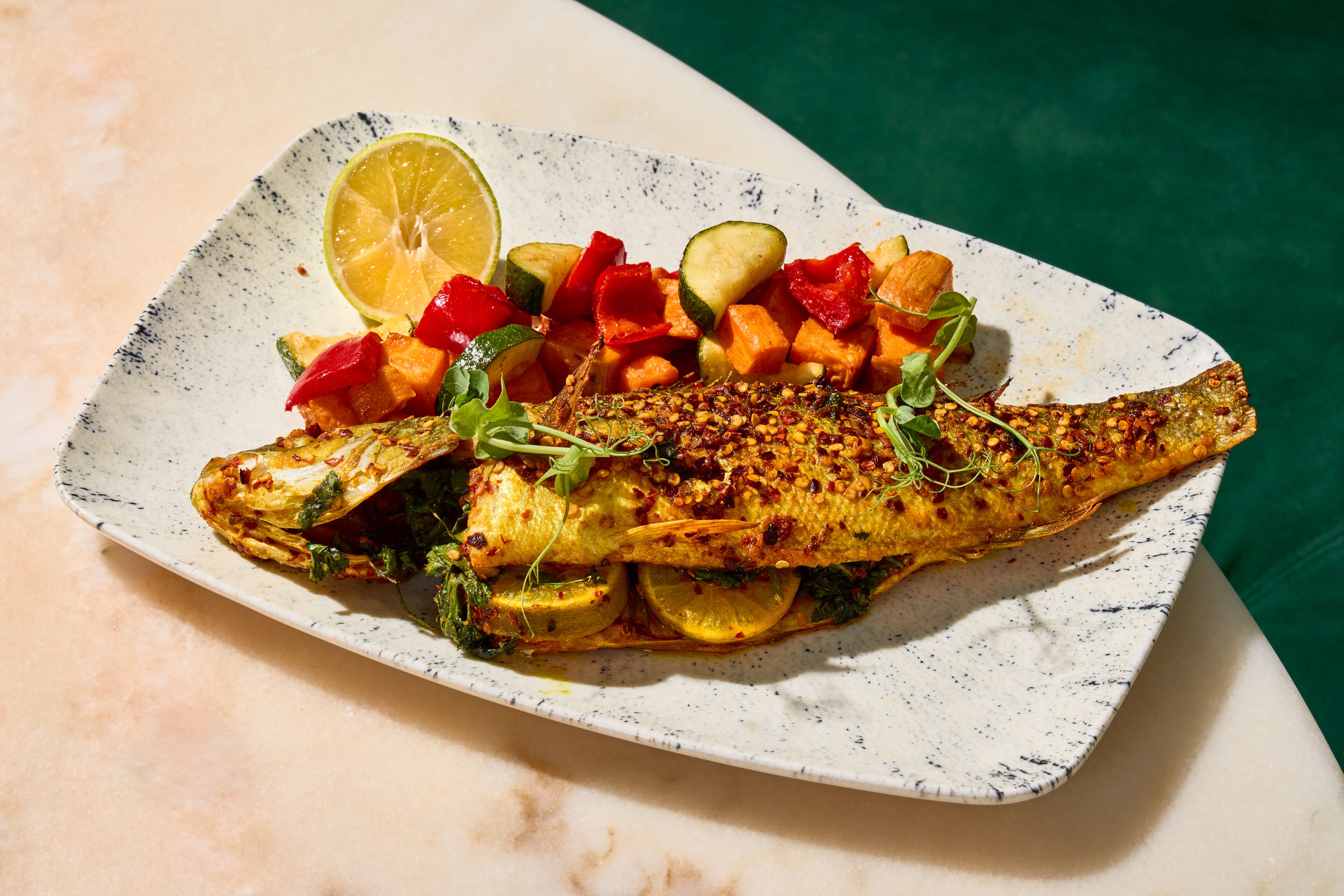 A plate of grilled fish with vegetables and a slice of lime.
