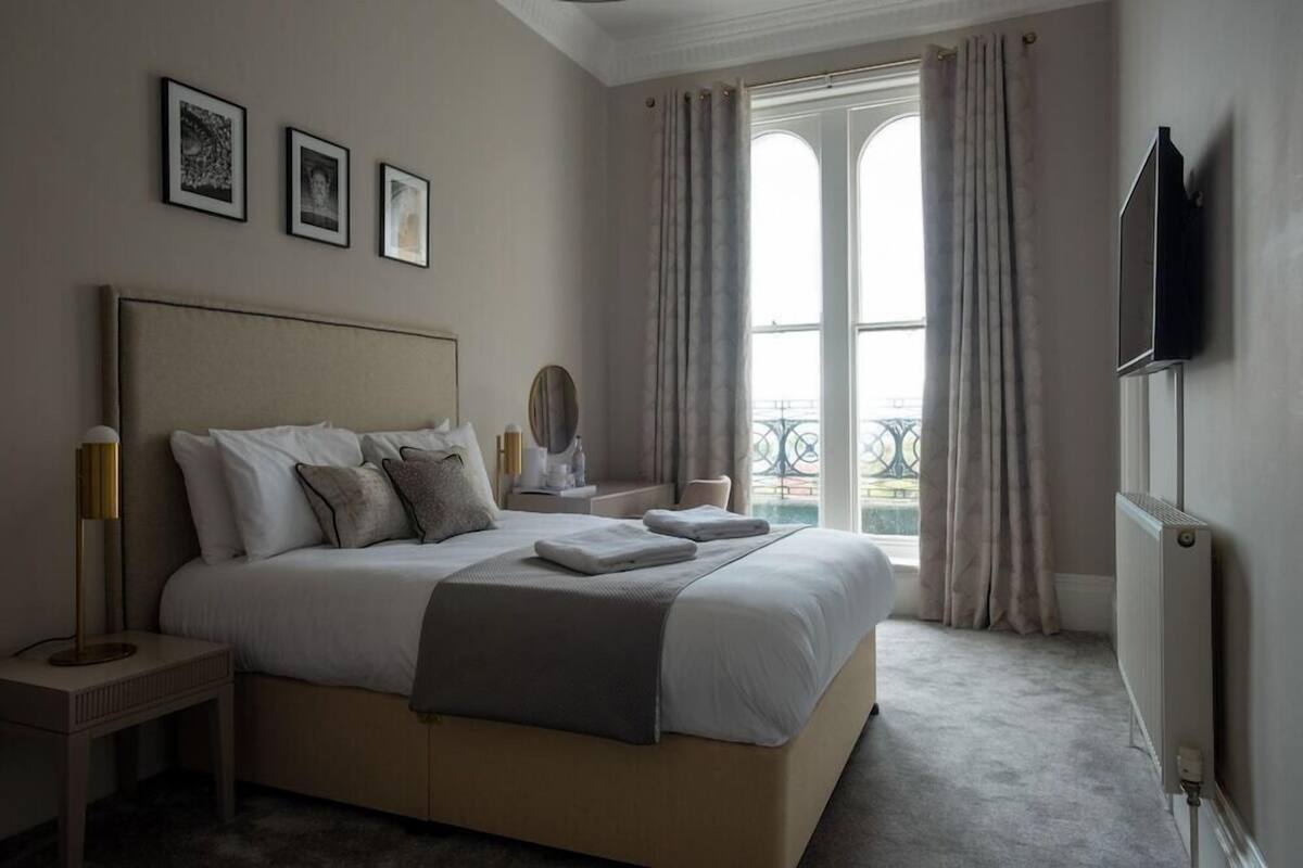 A bedroom with a large bed, a bedside table, a mirror, and a view of the outside through a window with curtains.
