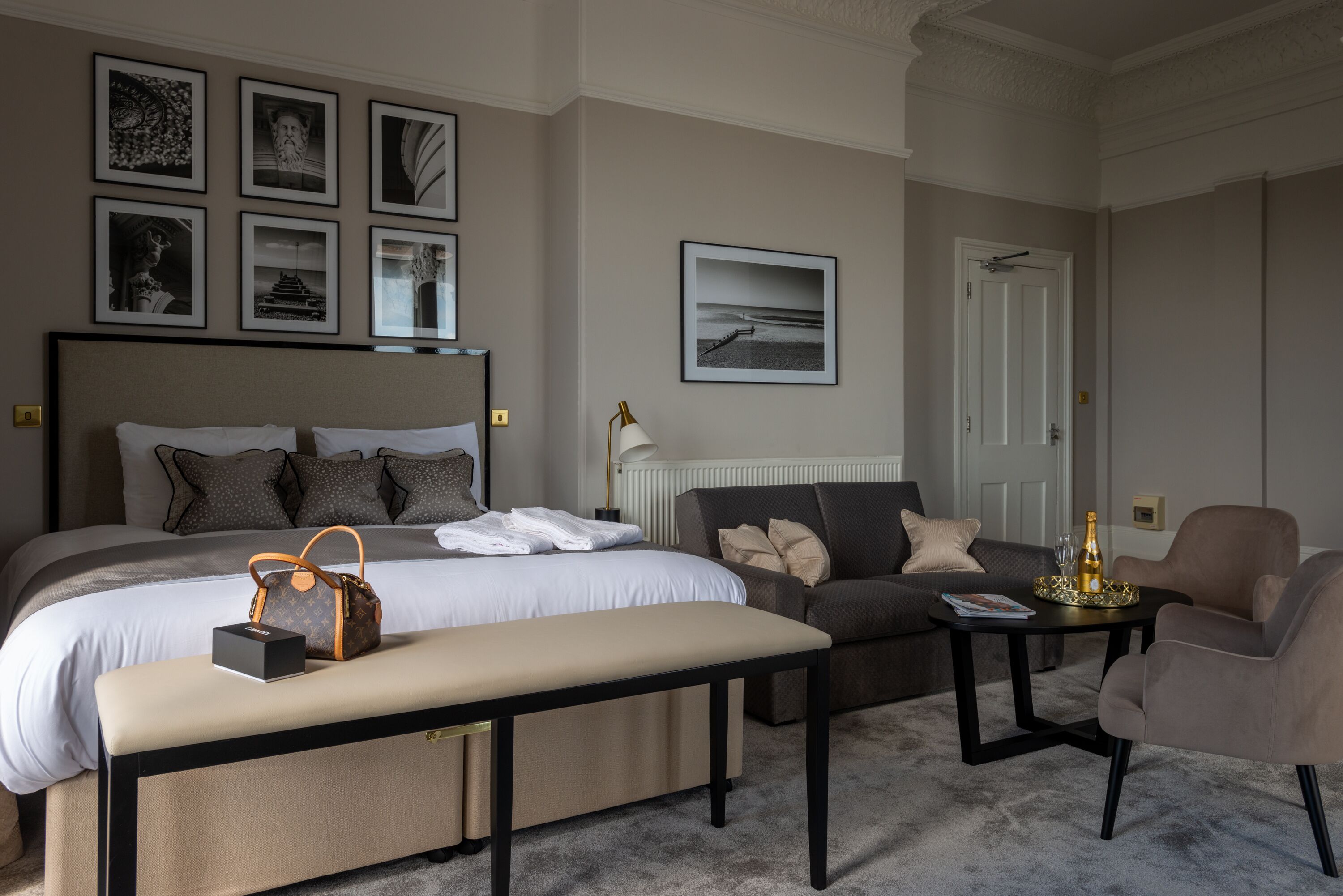 Habitación de hotel con una cama grande, un sofá, una mesita y un banco. Se observan cuadros en la pared y una puerta al fondo.