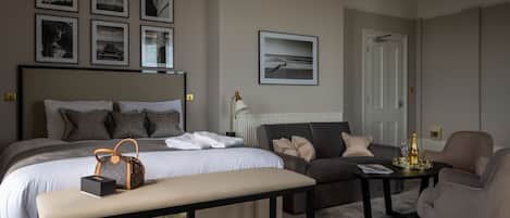 Habitación de hotel con una cama grande, un sofá, una mesita y un banco. Se observan cuadros en la pared y una puerta al fondo.