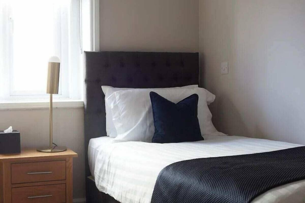 A neatly made bed with white and dark blue pillows, a wooden nightstand with a lamp, and a window with natural light.