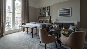 A modern hotel room with a bed, a sofa, a dining table, and a view of buildings through large windows. - The Burlington Hotel (Worthing)