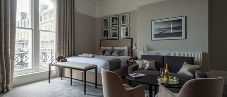 A modern hotel room with a bed, a sofa, a dining table, and a view of buildings through large windows.