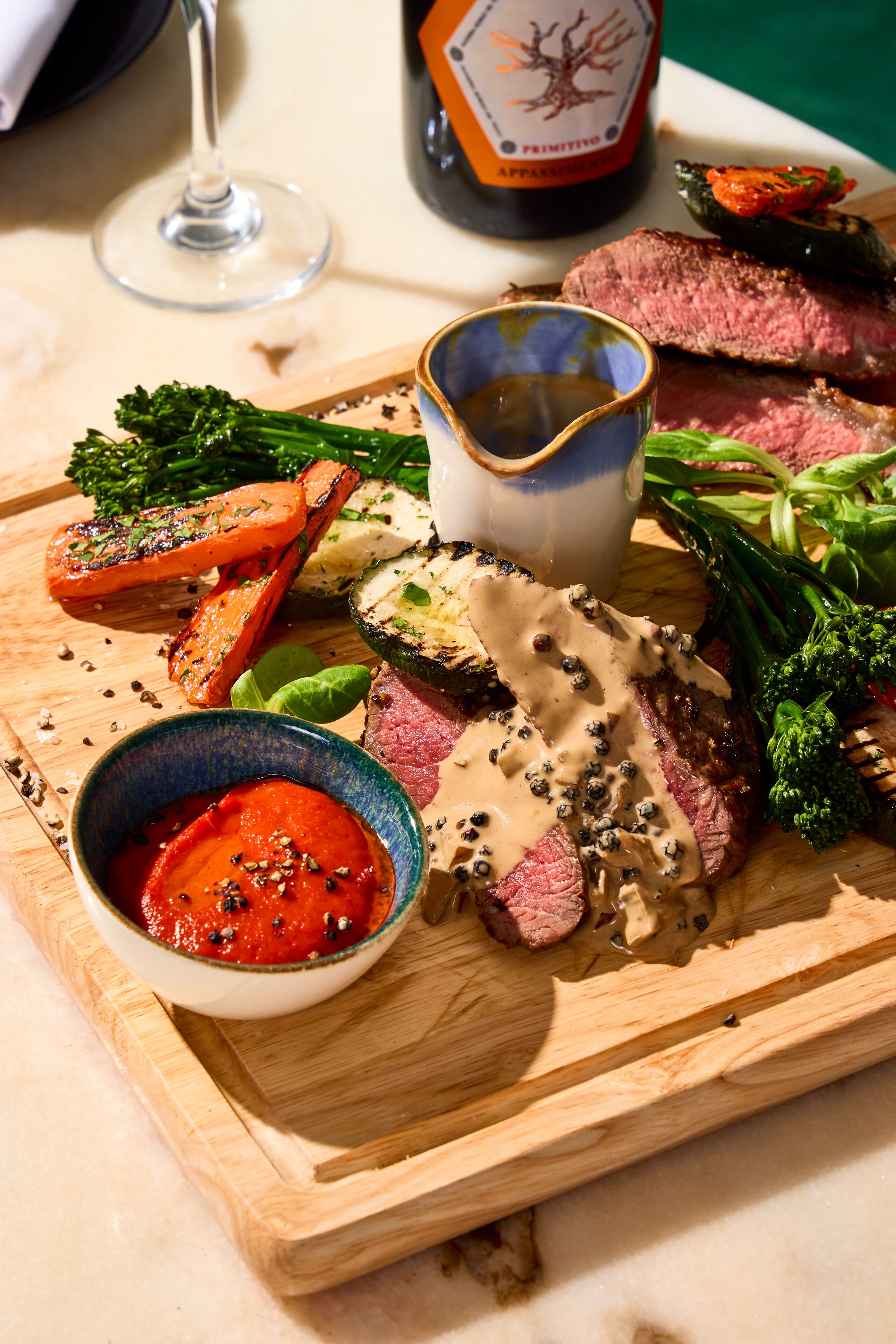 A wooden board with grilled vegetables, a bowl of sauce, and a bottle of wine.