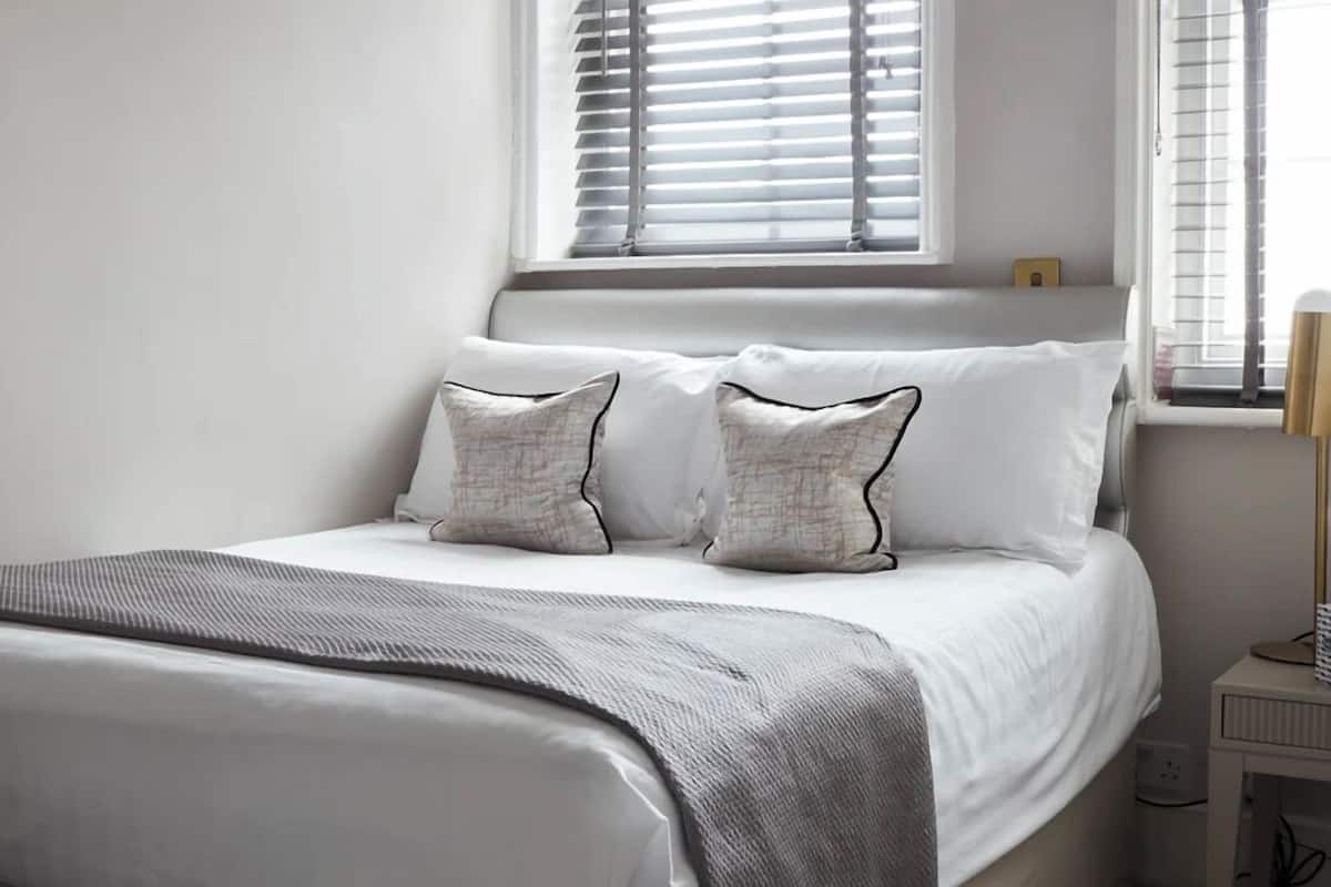 A neatly made bed with white linens and two patterned pillows, a bedside table with a lamp, and a window with blinds.