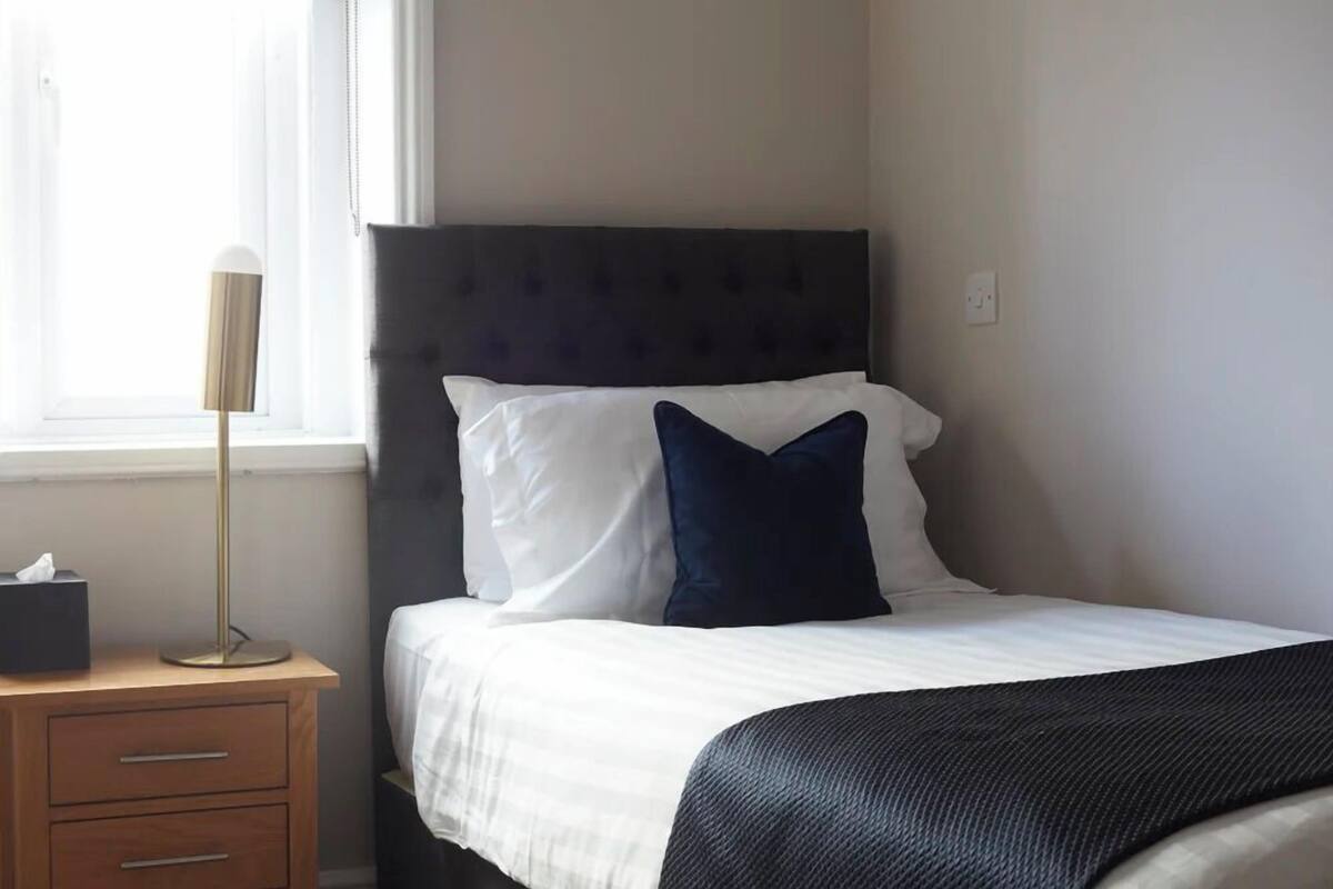 A neatly made bed with white and dark blue pillows, a dark headboard, and a bedside table with a lamp and a tissue box.