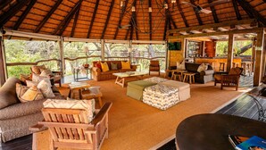 Un amplio espacio habitable de concepto abierto con un techo de paja alto, muebles de madera y una zona de bar visible a través de grandes ventanales.