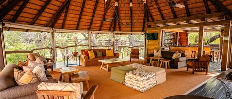 A spacious, open-plan living area with a high thatched ceiling, wooden furniture, and a bar area visible through large windows.