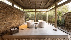 Un espacio de estar al aire libre cubierto con paredes de piedra, dos sillones acolchados, una mesita y una cesta de toallas.