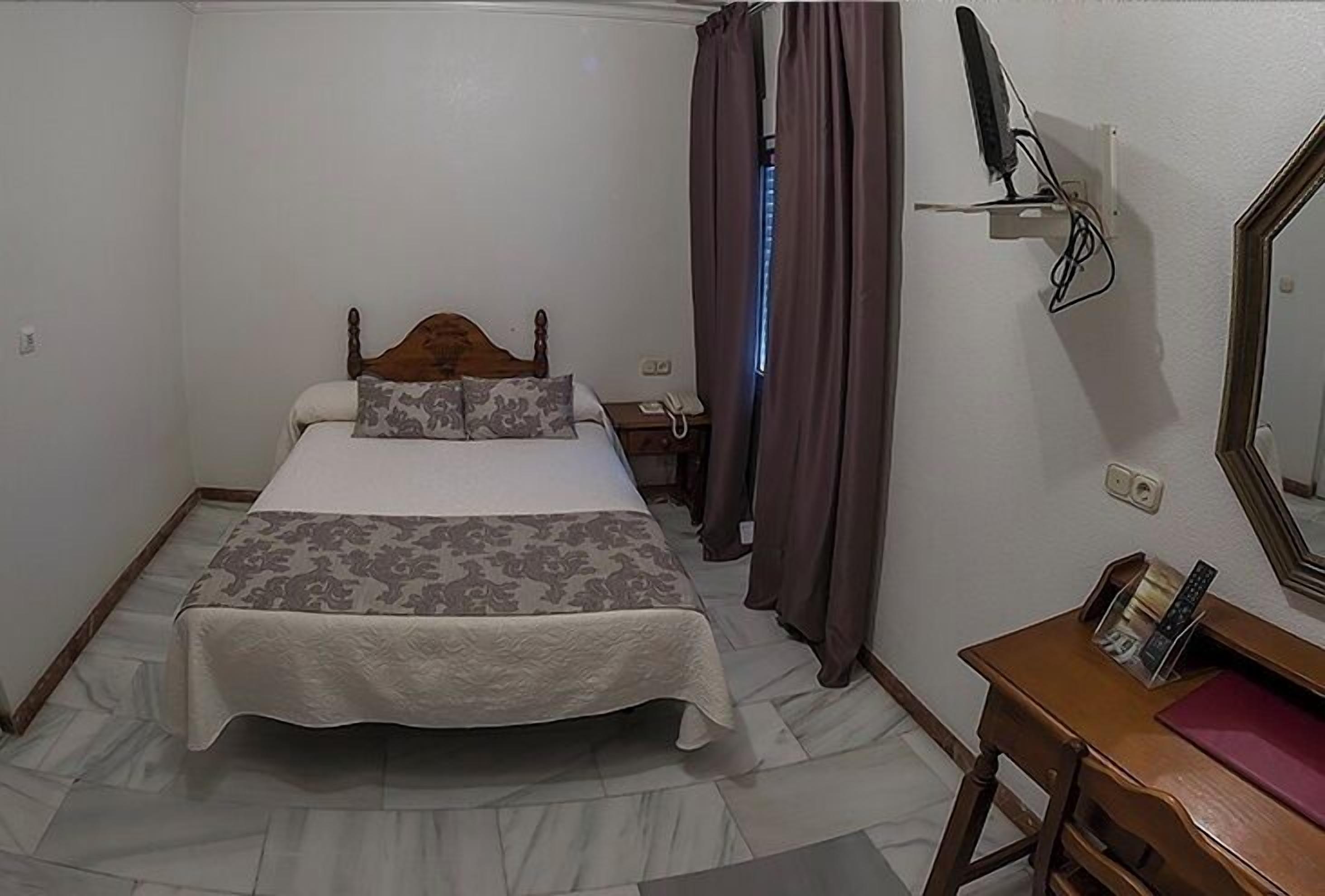 Un dormitorio con cama, cortinas, mesita de noche, espejo y televisión montada en la pared.