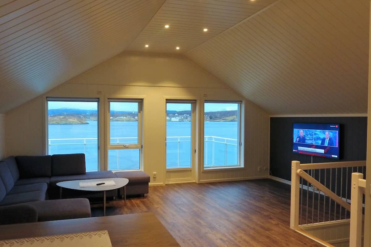 A cozy living room with a large window overlooking a body of water, a television on the wall, and a wooden ceiling.