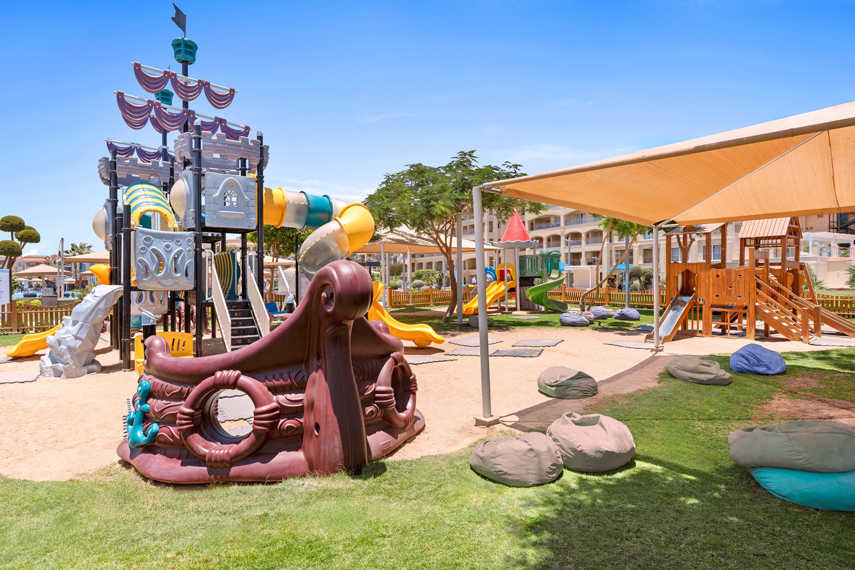A playground with slides and climbing structures, surrounded by a grassy area and a large umbrella providing shade.