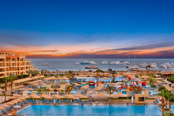 A resort with a large pool, multiple umbrellas, and a view of the sea with boats.