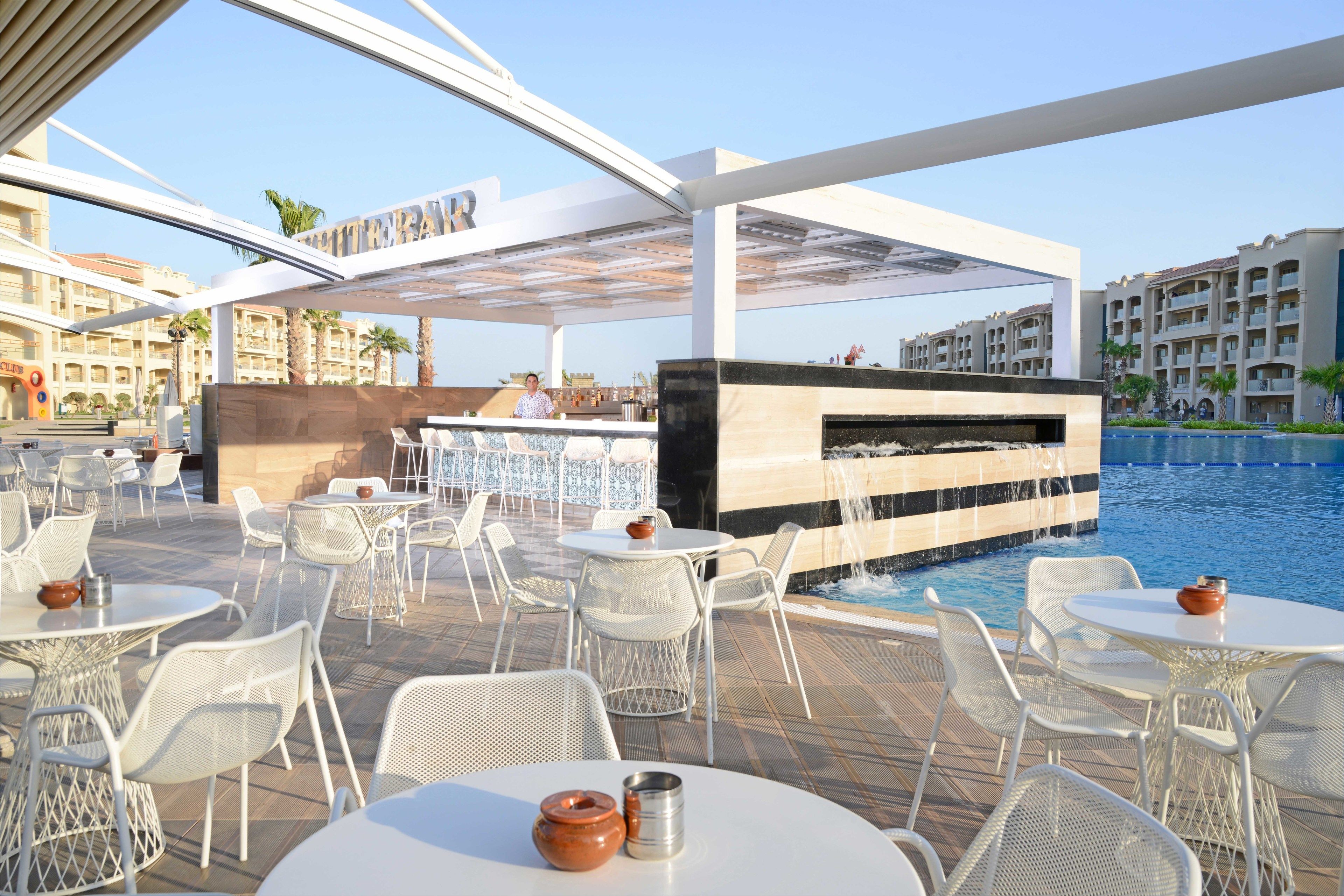 An outdoor pool area with white metal chairs and tables, a bar area, and a view of the pool and surrounding buildings.