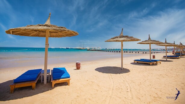 Ein Strand mit Liegestühlen und strohgedeckten Sonnenschirmen, ein Steg im Hintergrund und ein klarer, blauer Himmel.