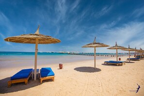 A beach with lounge chairs and thatched umbrellas, a pier in the background, and a clear blue sky.