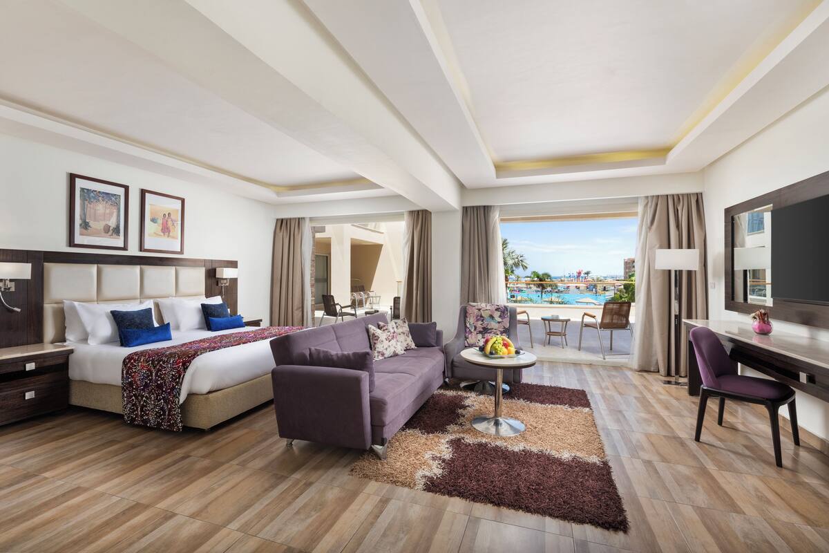 Een hotelkamer met een bed, bank, fauteuil, bureau en uitzicht op een zwembad.
