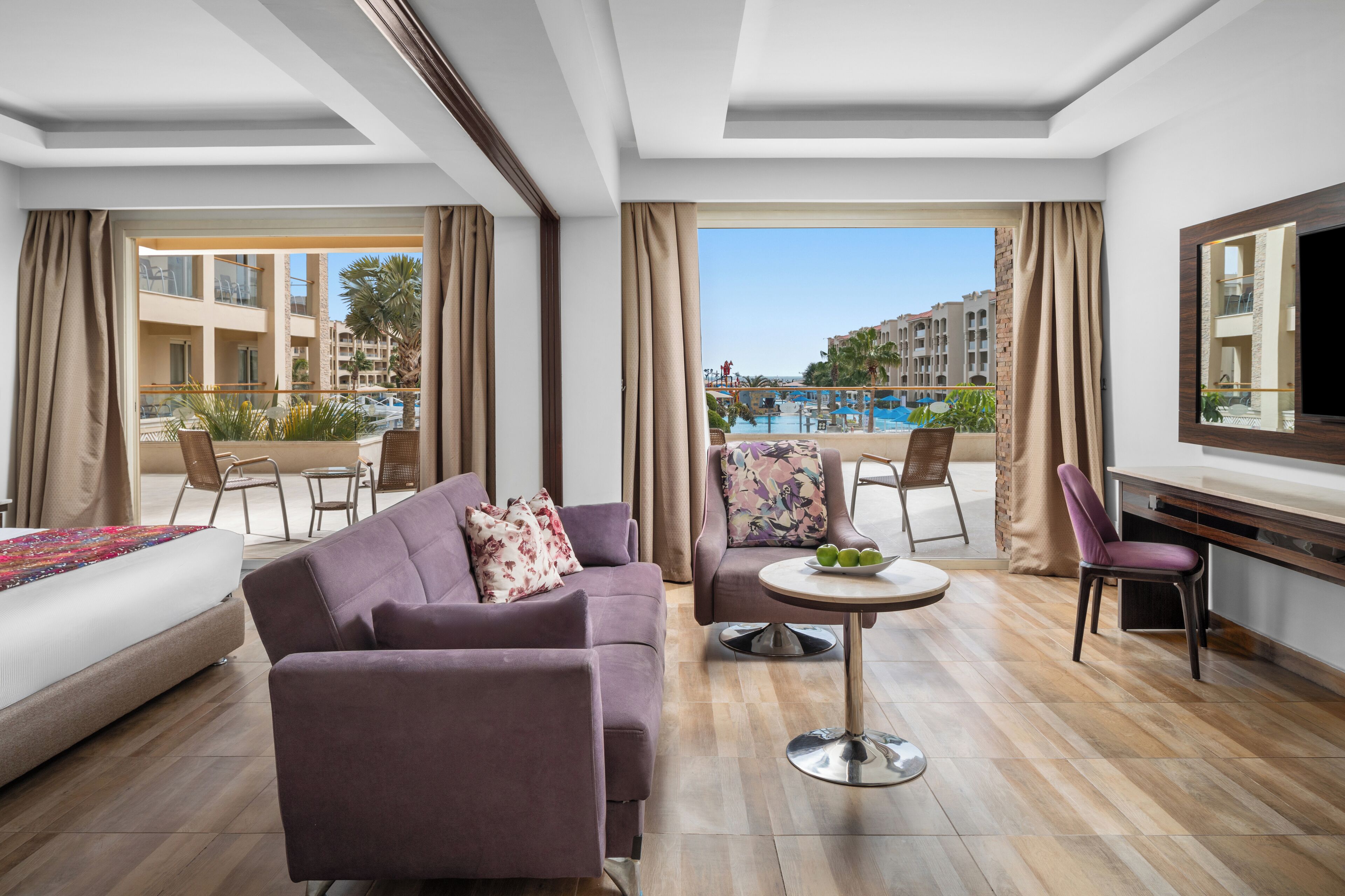 A hotel room with a large bed, a sofa, a dining table with chairs, and a view of a swimming pool and buildings.