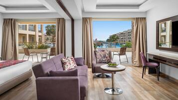 A hotel room with a large bed, a sofa, a dining table with chairs, and a view of a swimming pool and buildings.