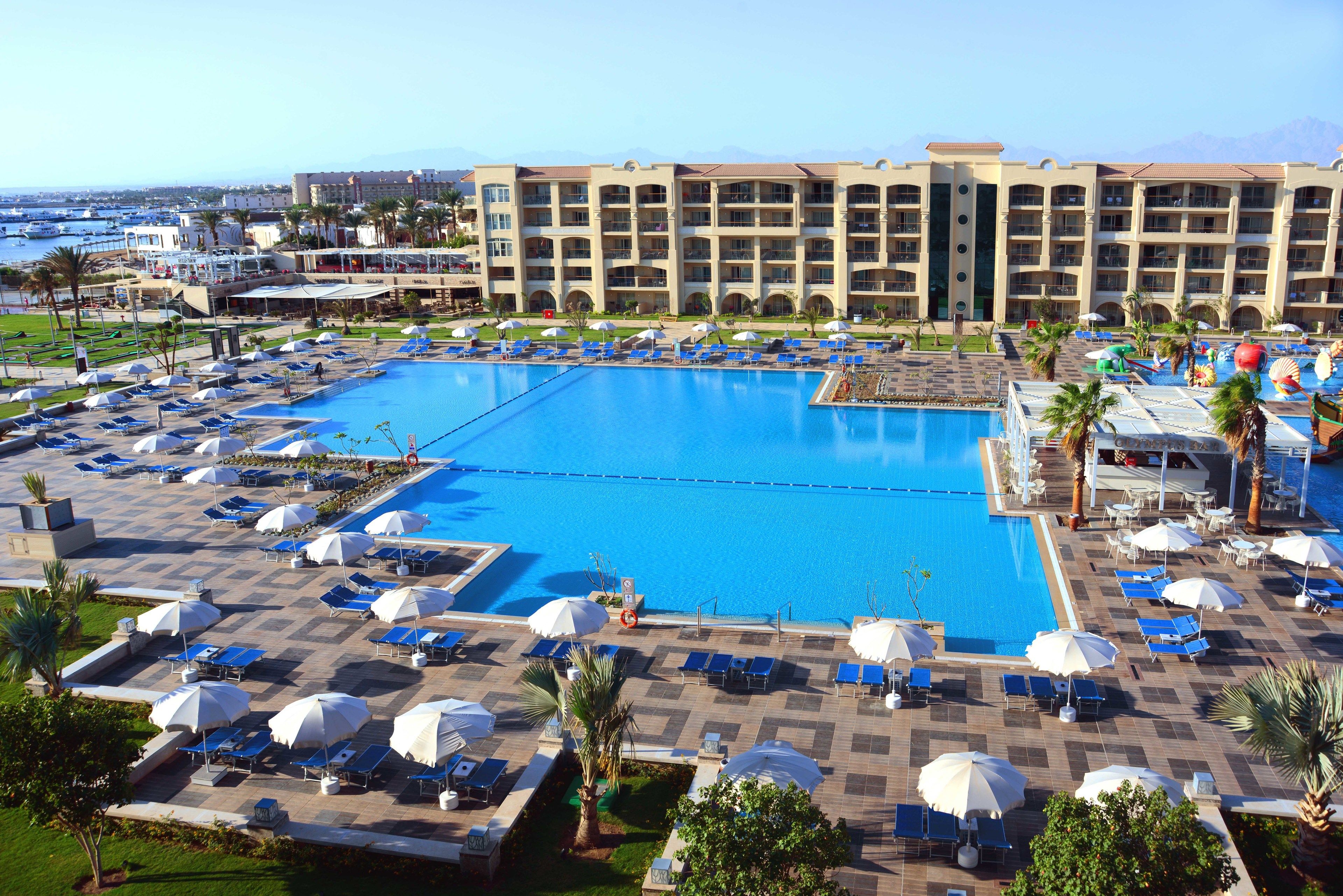 Una gran piscina con tumbonas y sombrillas, rodeada por un edificio de hotel con varios balcones.