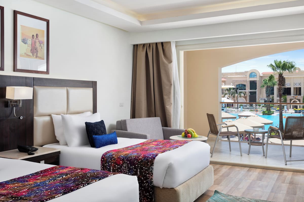 Hotelkamer met twee bedden, een bureau, een stoel, een kleine tafel met fruit, en uitzicht op een zwembad en palmbomen.