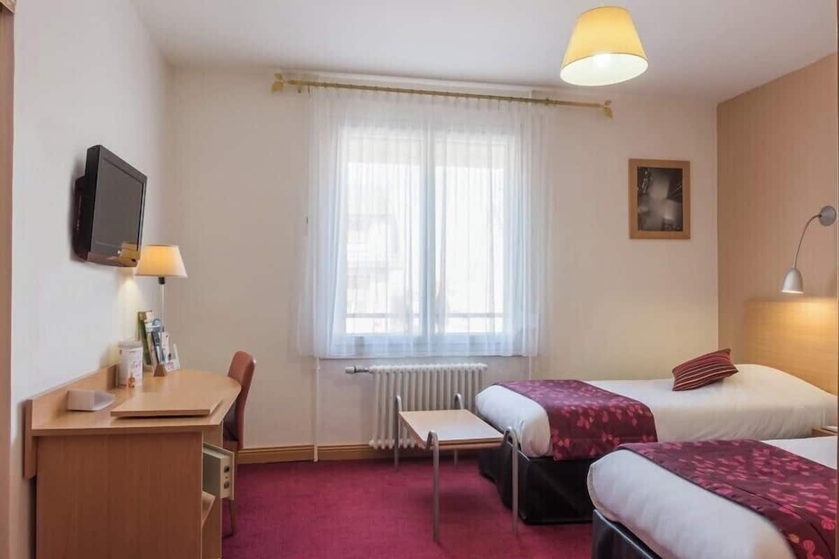 A hotel room with two beds, a desk, a chair, a radiator, a television, and a window with sheer curtains.