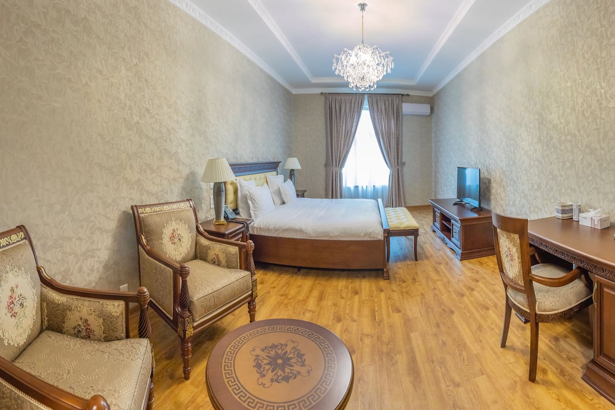 A hotel room with a bed, two armchairs, a desk, a television, and a chandelier.