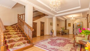 Una gran escalera con elegantes peldaños alfombrados, un candelabro y un tapete grande con motivos, en un amplio salón con suelo de madera y mobiliario clásico.
