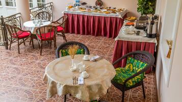 Un coin repas avec une table ronde dressée pour deux personnes, entourée de chaises en osier avec des coussins rouges. Un buffet, situé à l’arrière-plan, propose une variété de plats et de boissons.