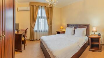 A hotel room with a large bed, a wooden wardrobe, a desk with a chair, a desk lamp, and a chandelier.
