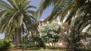 A garden with palm trees and flowering plants, a building with balconies, and a clear blue sky. - Globales Isabel (Son Servera)
