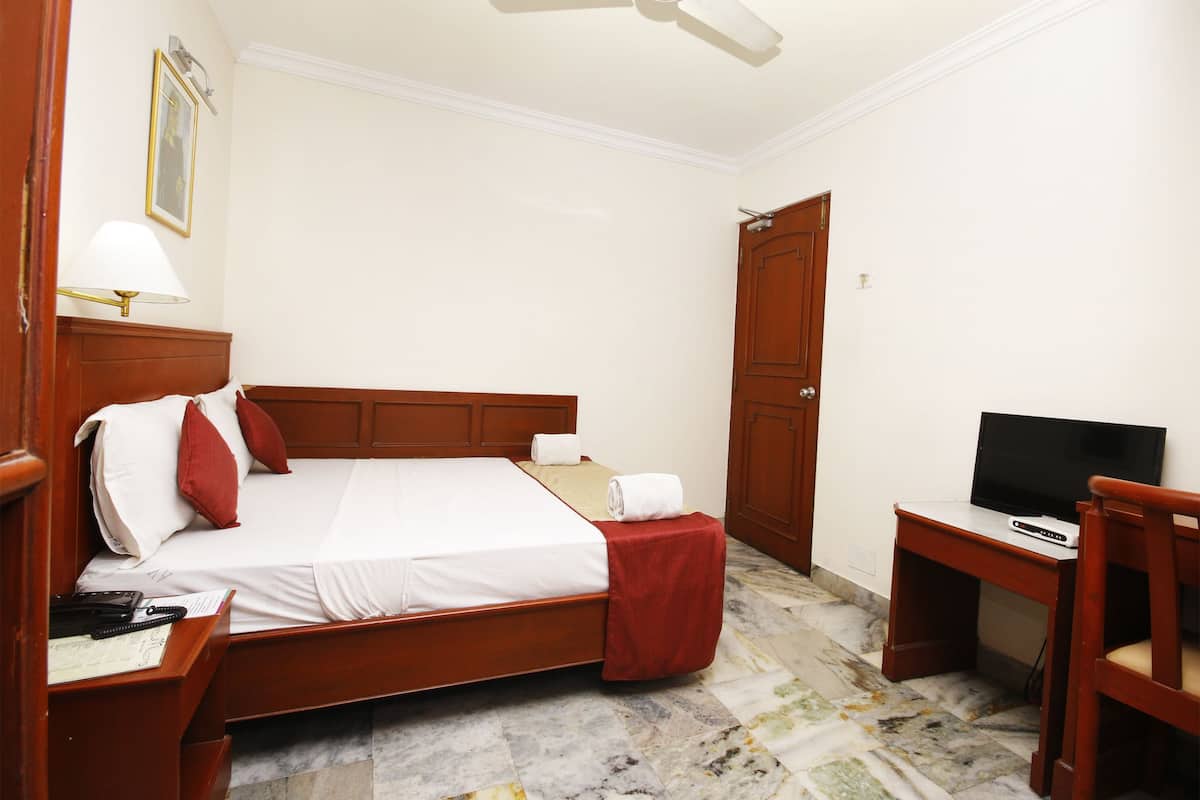 A hotel room with a bed, a desk with a television, a chair, and a wooden wardrobe.