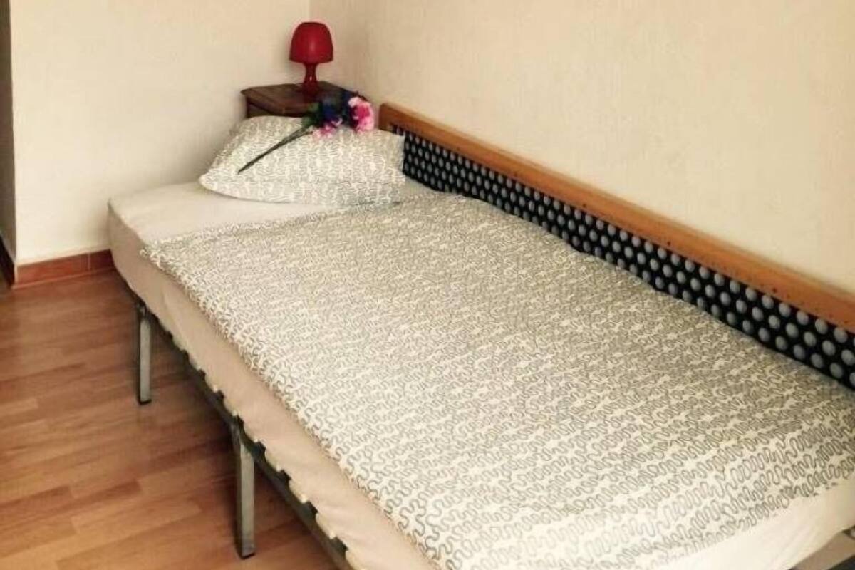 A small, single-bed room with a wooden headboard, a round mirror on the wall, and a small bedside table with a red lamp.