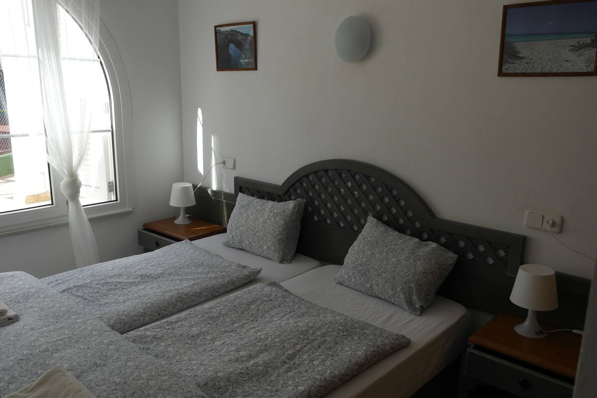 A bedroom with a bed, two bedside tables, a nightstand, a window with curtains, and wall-mounted artwork.