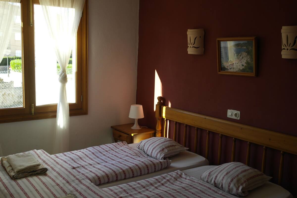 A bedroom with two beds, a nightstand with a lamp, a painting on the wall, and a window with sheer curtains.