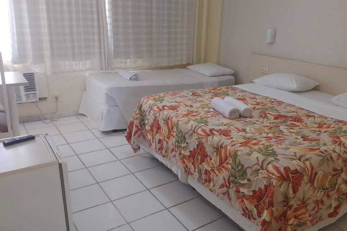 Ein Hotelzimmer mit zwei Betten, einem Schreibtisch mit Fernbedienung und einem Fenster mit Vorhängen.
