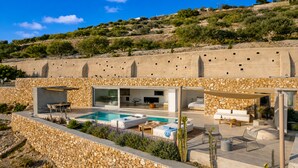 A modern villa with a swimming pool, outdoor seating, and a stone wall backdrop. - Santorini Heights (Santorini)