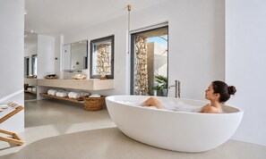 A modern bathroom with a freestanding bathtub, a large mirror, and a stone wall. - Santorini Heights (Santorini)