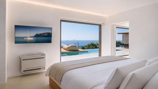 A bedroom with a bed, a wall-mounted air conditioner, a TV displaying a scenic view, and a large window overlooking a pool and outdoor seating area. - Santorini Heights (Santorini)